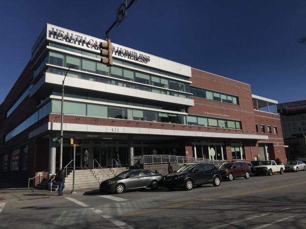 Großes Gebäude mit der Aufschrift "New York City Health Care Homeless Center" und parkenden Autos, Fußgängern auf einem Bürgersteig, Straßenmöblierung, Bäumen und einem klaren blauen Himmel.