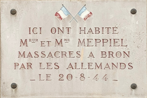 Steintafel an einem Gebäude mit zwei Fahnen darüber, die einen Text tragen, der ein Massaker gedenkt.