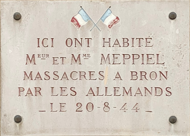 Steintafel an einem Gebäude mit zwei Fahnen darüber, die einen Text tragen, der ein Massaker gedenkt.