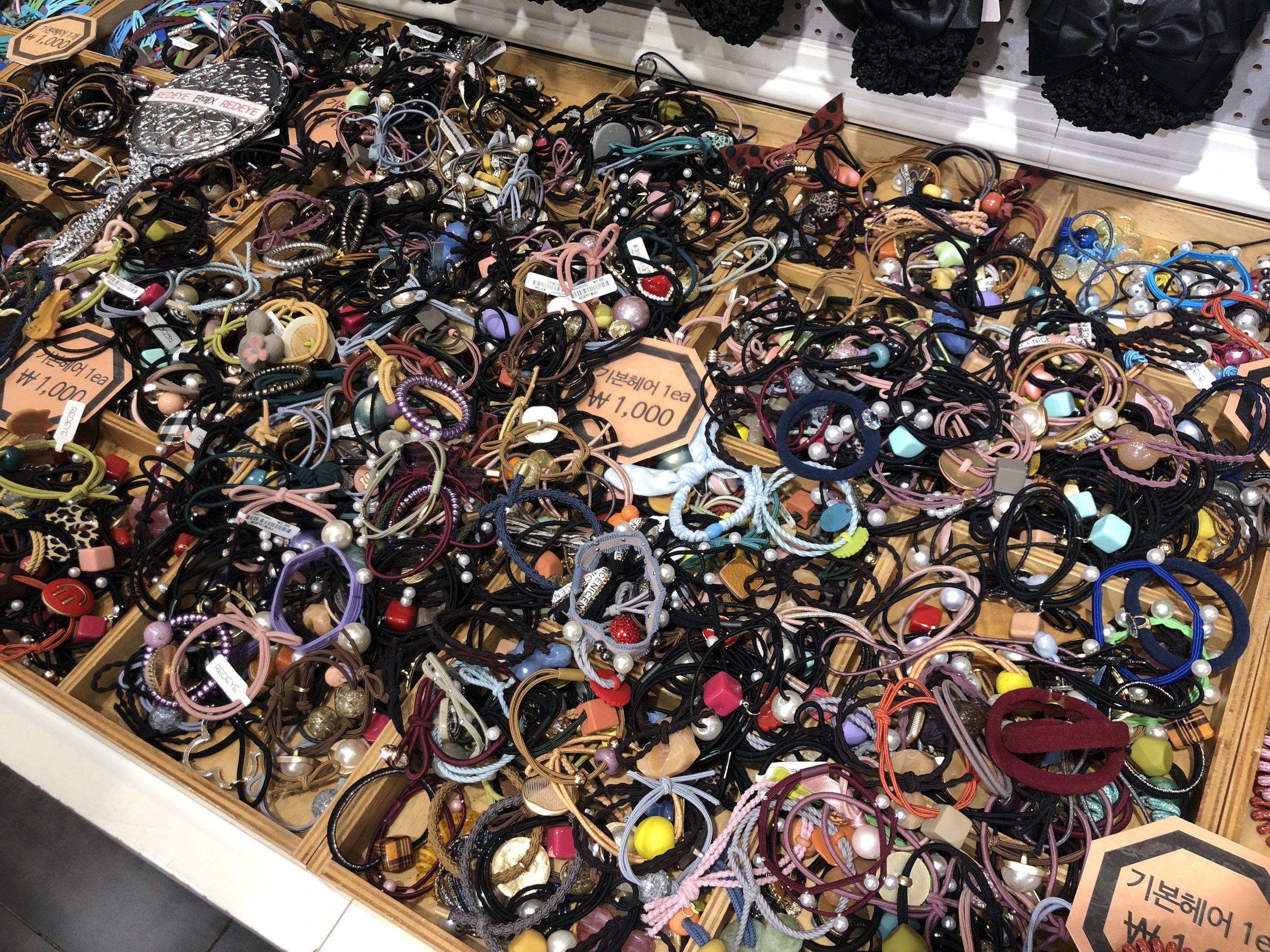 Vitrine voller verschiedener Schmuckstücke einschließlich Armbänder, Ketten und Preisschilder, mit Kleidung darüber, die einen Marktplatz suggeriert.