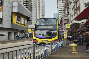 Ein gelber Doppeldeckerbus fährt eine Straße mit hohen Gebäuden entlang, mit Fußgängern auf dem rechten Gehweg, einem Hydranten und einem Geländer, unter einem klaren blauen Himmel.