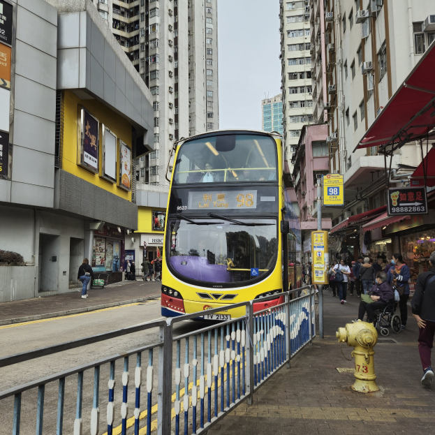 Ein gelber Doppeldeckerbus fährt eine Straße mit hohen Gebäuden entlang, mit Fußgängern auf dem rechten Gehweg, einem Hydranten und einem Geländer, unter einem klaren blauen Himmel.