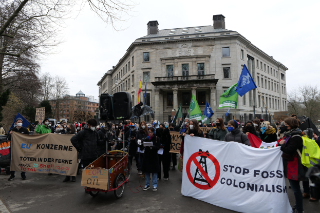 Large group of people marching in a protest against fossil fuels, carrying banners and flags, with a vehicle in the foreground and buildings, trees, and a clear blue sky in the background.