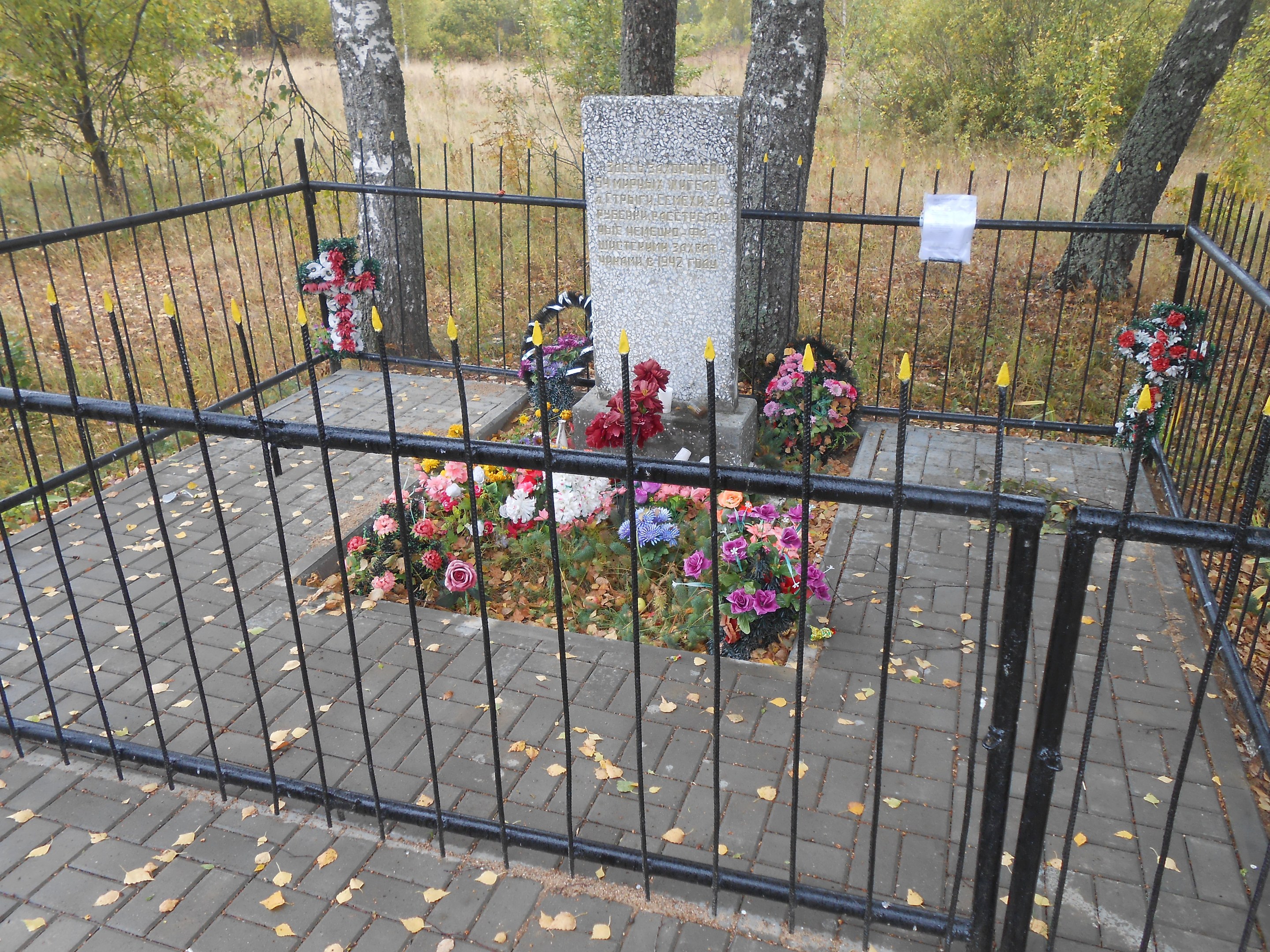 Ein von einem Metallzaun umgebenes Denkmal mit Blumensträußen in einem Park mit Bäumen, Gras und trockenen Blättern auf dem Boden.