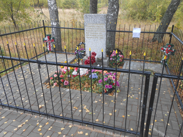 Ein von einem Metallzaun umgebenes Denkmal mit Blumensträußen in einem Park mit Bäumen, Gras und trockenen Blättern auf dem Boden.