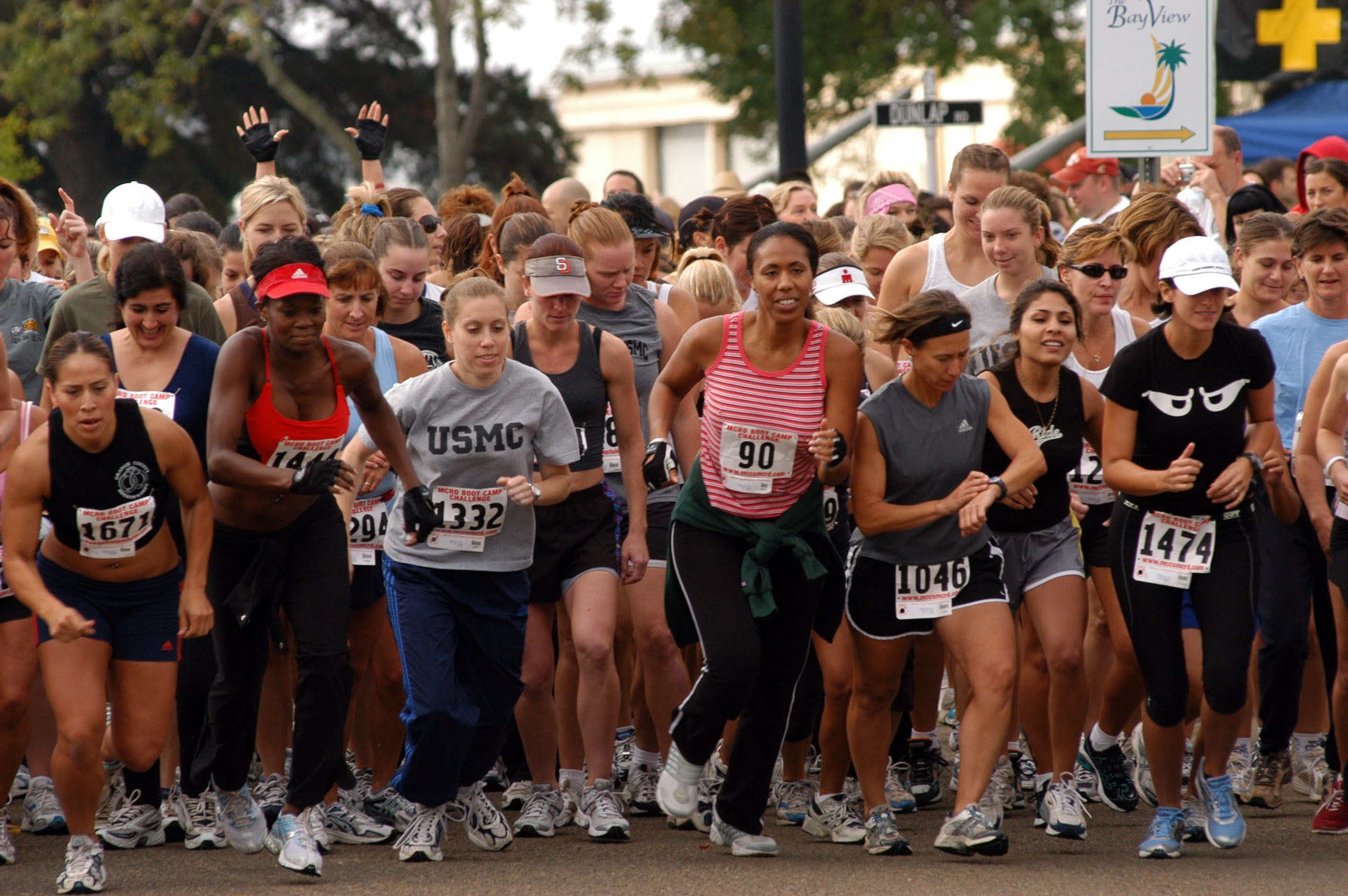 Gruppe von Frauen beim Marathon auf einer Straße mit Bäumen, Gebäuden, Schildern, Zelten und anderen Objekten im Hintergrund, aufgenommen mit Bewegungsunschärfe.