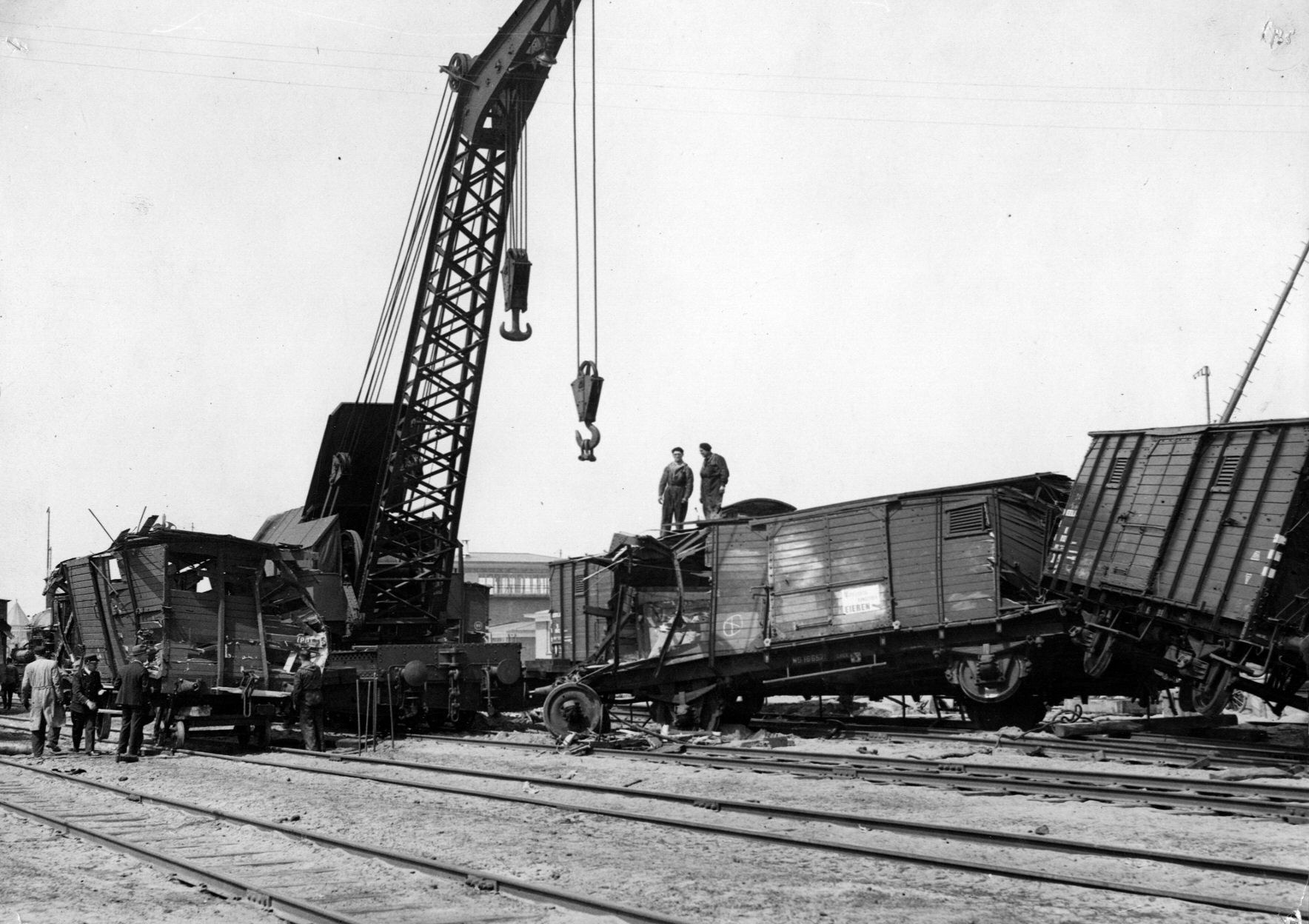 Schwarzes und weißes Bild einer Zugkatastrophe mit einem Kran, der einen Zugwagen auf die Schienen hebt, mehrere Menschen in der Nähe, Gebäude und ein klarer Himmel im Hintergrund.