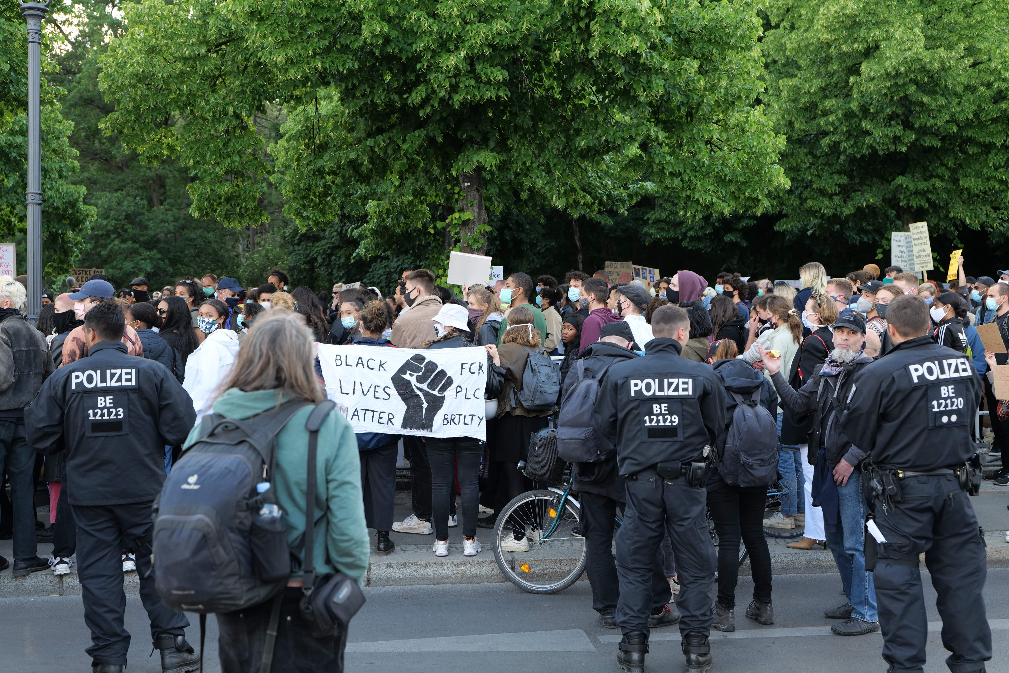 Eine große Gruppe von Menschen nimmt an einem Black Lives Matter-Protest auf der Straße teil, einige halten Schilder und andere tragen Kappen und Taschen, mit einem Fahrrad im Vordergrund und Bäumen und einem Mast im Hintergrund.