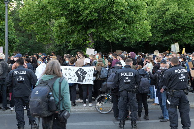 Eine große Gruppe von Menschen nimmt an einem Black Lives Matter-Protest auf der Straße teil, einige halten Schilder und andere tragen Kappen und Taschen, mit einem Fahrrad im Vordergrund und Bäumen und einem Mast im Hintergrund.