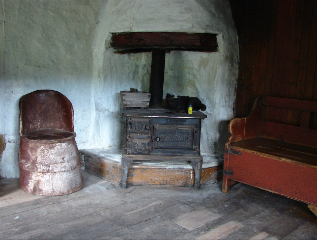 Alte Holzofen in einem Raum mit einer hölzernen Bank auf der rechten Seite, einem Stuhl auf der linken Seite und einer Wand im Hintergrund.