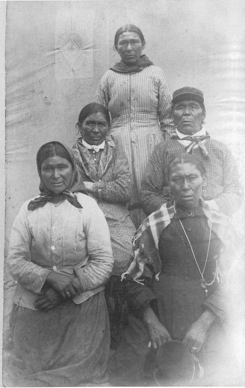 Schwarz-weiß-Foto einer Gruppe von indigenen Frauen, die in einer Reihe vor einer Wand stehen, einige tragen traditionelle Kleidung und Kopfschmuck.