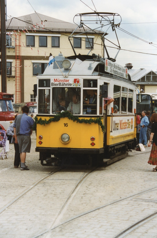 Ein gelb-weißer Straßenbahnwagen auf einer Stadtstraße mit Menschen zu seiner Rechten, Gebäuden, Polen, Drähten und einem klaren blauen Himmel im Hintergrund.