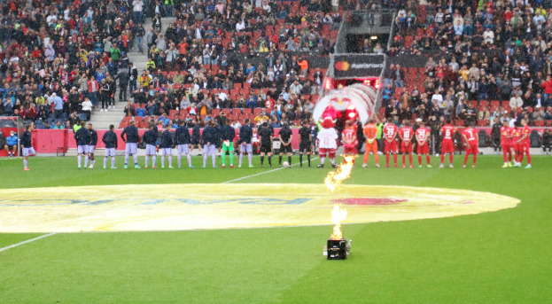 Gruppe von Menschen auf einem Fußballfeld mit einem Feuer in der Mitte, während andere auf Stühlen, Treppen, Geländern und anderen Gegenständen im Hintergrund sitzen.