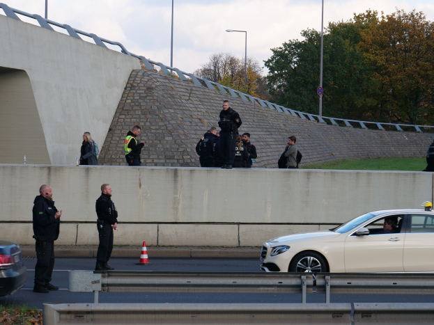 Police officers standing on the side of a road next to a car with traffic cones, a divider, grass, a wall, light poles, trees, and a cloudy sky in the background.