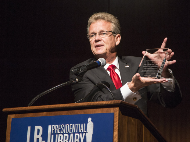 Ein Mann in Anzug und Krawatte mit einer Auszeichnung auf einem Podium mit einem Mikrofon und einer Tafel, vor einem dunklen Hintergrund bei einer Bibliothek-Veranstaltung.