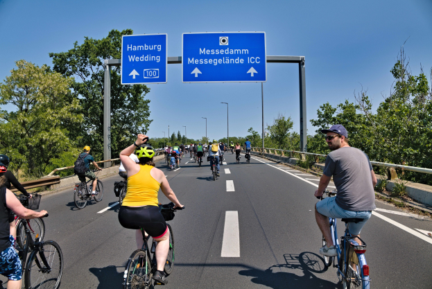 Group of people wearing helmets riding bicycles down a road with a railing on one side and trees on the other, light poles in the background, and a clear blue sky.