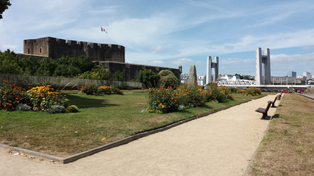 Park mit Bänken und bunten Blumen vor einer Burg, umgeben von Grün, einem Zaun, Bäumen, Fahrzeugen, einer Brücke, Säulen, einem Fahnenmast mit Flagge, Gebäuden und einem bewölkten Himmel.