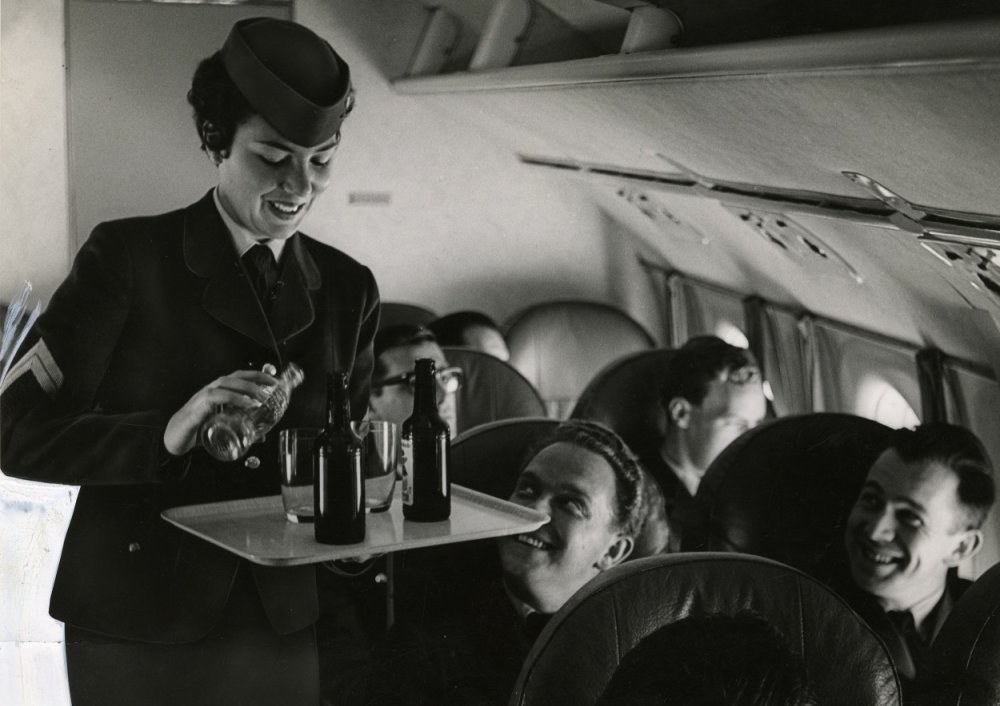 Schwarzes und weißes Bild einer Flugbegleiterin, die Getränke an sitzende Passagiere auf einem Flugzeug serviert.