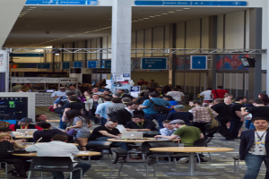 Ein belebter Flughafenempfang mit wartenden Menschen, einige stehen und arbeiten, Richtungsschilder an den Wänden und an der Decke sowie Deckenleuchten.