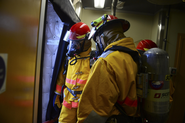 Feuerwehrleute in gelben Uniformen und roten Helmen stehen in einer Gruppe rechts neben Zylindern und links neben einer Tür, mit einer Wand und Licht im Hintergrund.