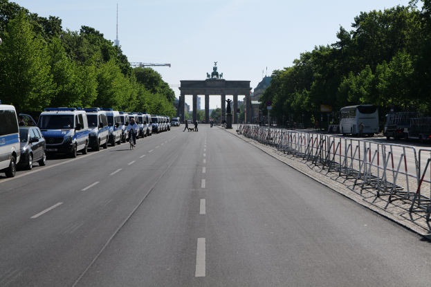 Eine lange Reihe von Polizeiwagen, die auf der Seite einer Straße vor dem Brandenburger Tor in Berlin, Deutschland, geparkt sind, mit Menschen, die Fahrräder fahren und auf der Straße, Barrieren und Bäume an den Seiten stehen, und ein Bogen mit Statuen im Hintergrund.