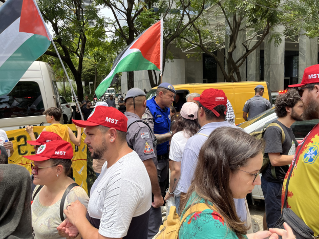 Eine Gruppe von Menschen steht vor einem Gebäude während einer Demonstration, einige tragen Mützen und Taschen, mit Fahrzeugen auf der Straße, Bäumen und einem klaren blauen Himmel im Hintergrund; mehrere halten palästinensische Flaggen.