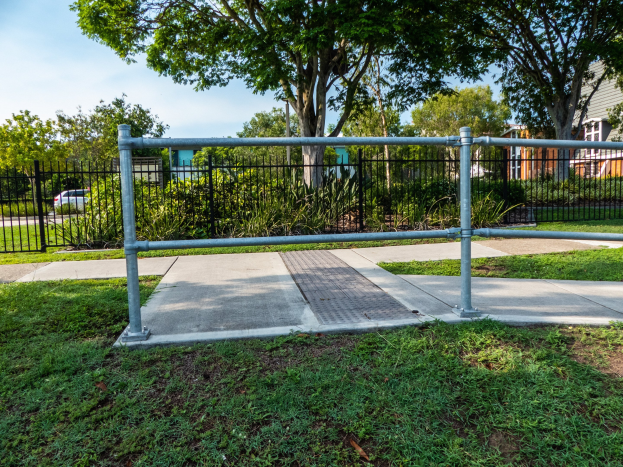 Metallzaun in einer grasbewachsenen Area mit umliegenden Pflanzen, Bäumen, einem Auto auf der Straße, Gebäuden im Hintergrund und einem klaren blauen Himmel.