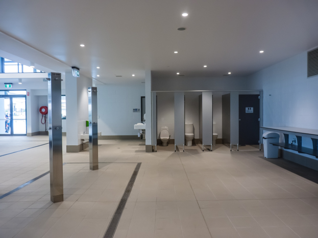 Großes öffentliches WC mit mehreren Toiletten, tragenden Säulen, einem Müllbehälter, einer Tür, einem Schild, einem angebrachten Feuerlöscher und Deckenleuchten.