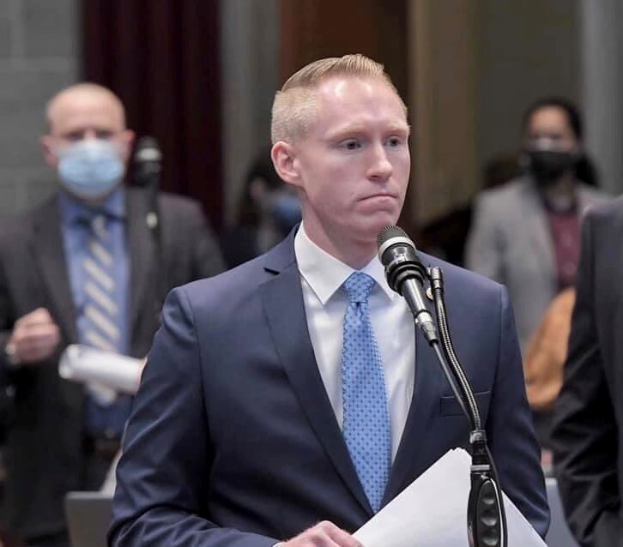 Ein Mann in Anzug und Krawatte spricht an einem Rednerpult mit Papieren, während andere in der Nähe stehen und eine Vorhangwand im Hintergrund zu sehen ist.