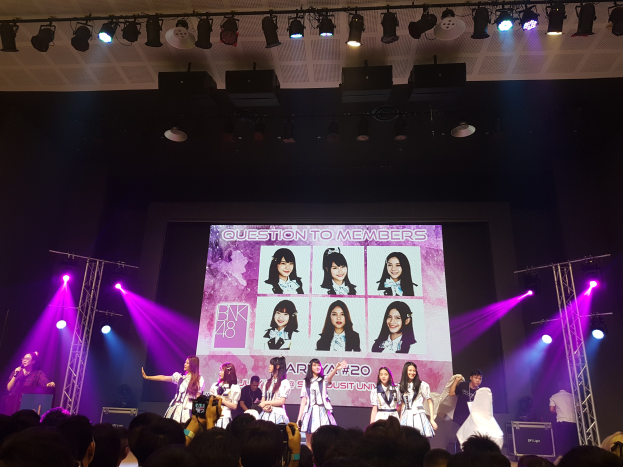 Eine Gruppe von Menschen performs auf einer hell erleuchteten Bühne mit Trägern, während ein Sänger ein Mikrofon hält und ein anderer ein Instrument spielt; unten schaut eine Menge mit Kameras zu, während ein großer Bildschirm im Hintergrund Bilder der K-Pop-Gruppe BK48 anzeigt.