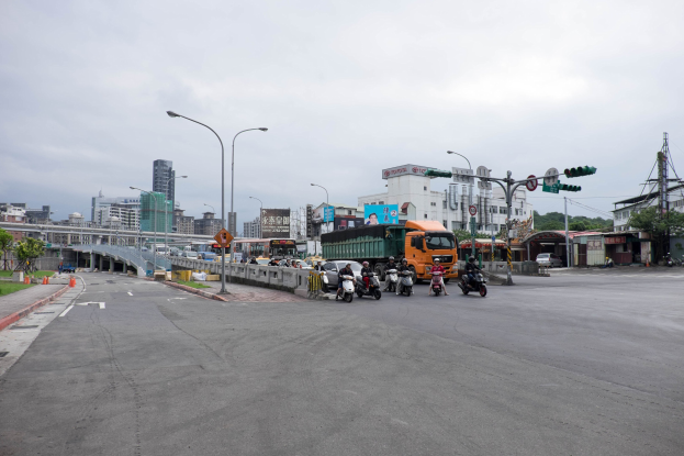 A city street with heavy traffic, including a truck and a car, featuring light poles, traffic signals, signboards, a bridge with railings, trees, buildings with windows, and a cloudy sky.