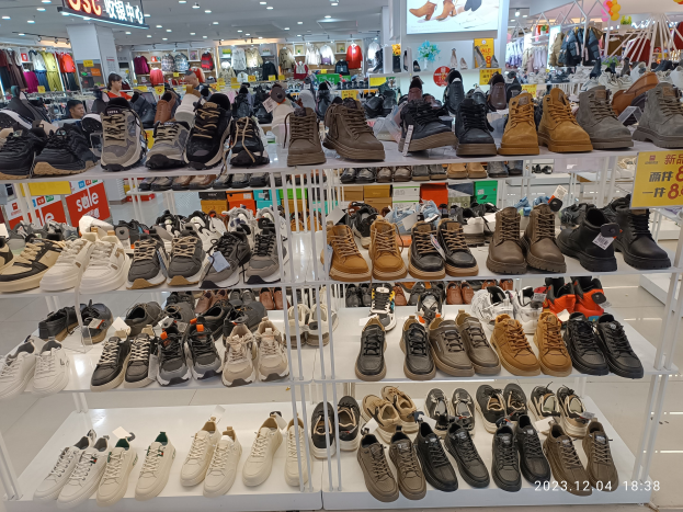 Ein Schuhgeschäfts-Innenraum mit verschiedenen Schuhen auf Gestellen, Preisetiketten sichtbar, Menschen im Hintergrund, Kleidung an Bügeln, Deckenleuchten, ein Bildschirm und Texttafeln, mit einem Wasserzeichen, das einen Laden in China anzeigt.