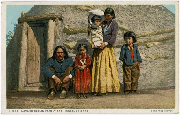 Black-and-white postcard of a Navajo family posing in front of a wooden house with a door, trees, and a clear sky, labeled "Navajo Indian Family and Hogan, Arizona" at the bottom.