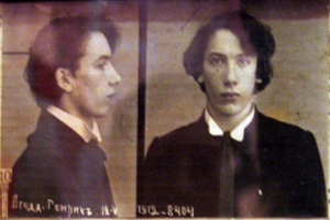 Ein Fahndungsfoto eines Mannes in einem schwarzen Anzug, weißem Hemd und schwarzer Krawatte mit ernster Miene, geföhnten Haaren und verschränkten Händen, begleitet von zwei zusätzlichen Fotos und fettgedrucktem Text über seine Verbrechen.