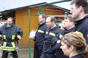 Eine Gruppe von Männern in blauen Feuerwehreinsatzuniformen steht nach links gewandt, mit einem kleinen Holzhäuschen und Rohrrahmen im Hintergrund.