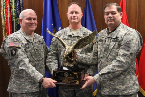Drei Männer in Uniformen stehen vor Flaggen, jeder hält eine Auszeichnung, mit bunten Elementen und Text in der oberen linken Ecke.