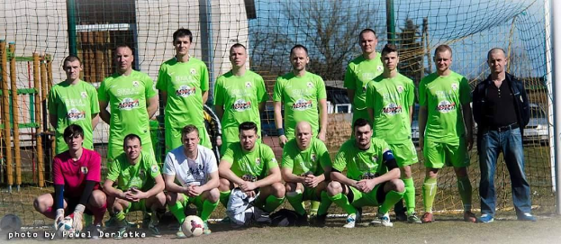 Group of men in green t-shirts standing on a soccer field with a ball and goal post, trees and buildings in the background.