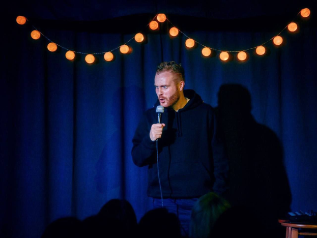 A man stands on a brightly lit stage holding a microphone, with a table of objects to his right and a curtain in the background, as the heads of an audience are visible below.