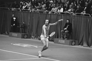 Ein Mann in Aktion schwingt einen Tennisracket auf einen Ball auf einem Court, mit zwei sitzenden Individuen und Vorh├Ąngen im Hintergrund, dargestellt in Schwarz-Wei├č.