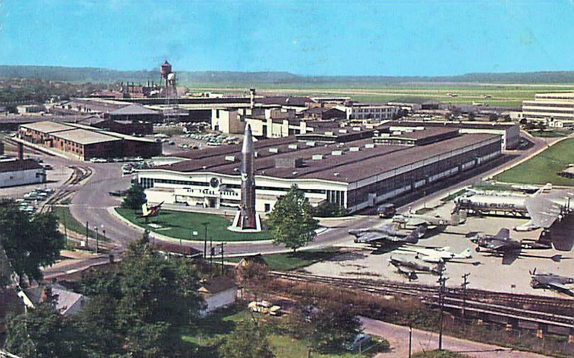 Altes Schwarz-Weiß-Foto eines Flughafens mit geparkten Flugzeugen, Gebäuden, Bäumen, Straßen, Fahrzeugen, Strommasten, Gras, Bahnschienen, Türmen, Hügeln und einem klaren blauen Himmel, mit Text unten.