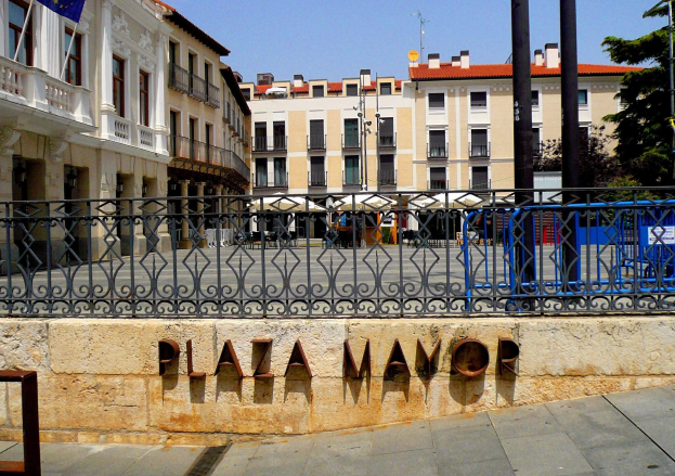 Plaza Mayor in Madrid, Spanien, mit einer Wand mit Text, einem Metallzaun, Gebäuden mit Fenstern, Pfählen, einem Schild, Bäumen und einem bewölkten Himmel.