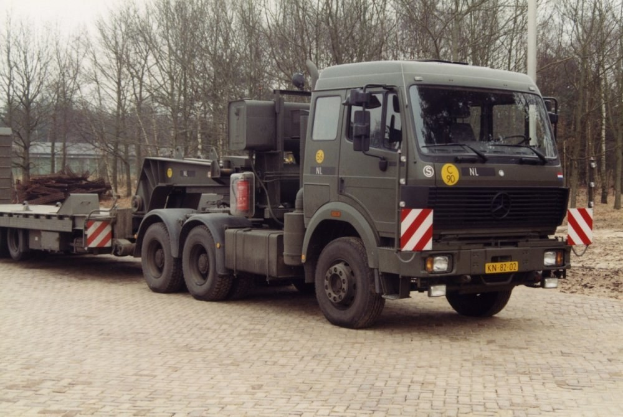 Ein großer Lastwagen mit einem Sattelauflieger parkt vor einem Gebäude, umgeben von Bäumen unter einem klaren blauen Himmel, mit einem sichtbaren Vorderrad.