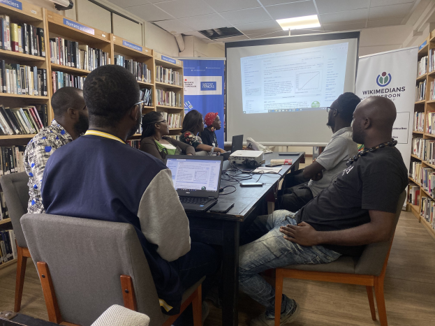 Gruppe von Menschen, die um einen Tisch mit Laptops und Kabeln sitzen und an einem Workshop zur Technologie in der Bildung teilnehmen, mit Projektorwand, Bannern, Bücherregalen und Deckenbeleuchtung.