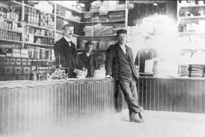 Drei Männer stehen an einem Tresen in einem Laden mit verschiedenen Gegenständen, ein Buchregal im Hintergrund, scheinbar mitten in einem Raubüberfall.