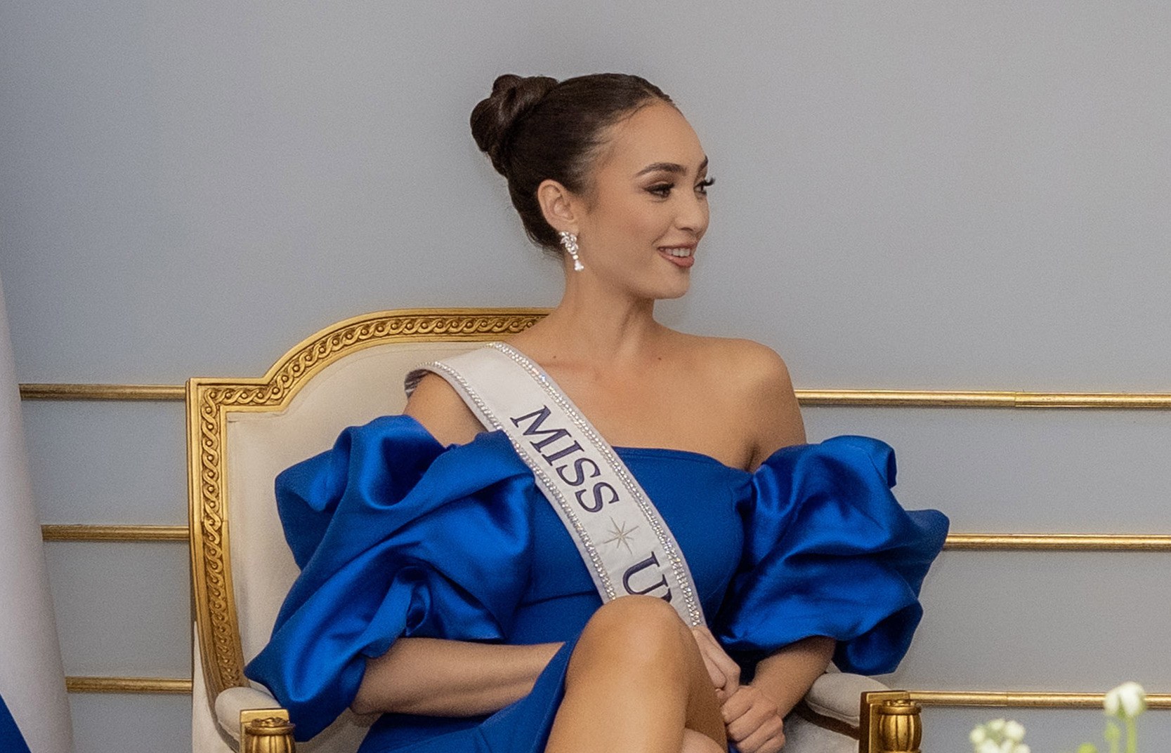 Frau in einem blauen Kleid mit einem "Miss Universe 2019"-Band, die auf einem Stuhl sitzt und von Blumen umgeben ist.