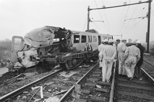 Ein Schwarz-Weiß-Bild einer Zugkollision mit einer Gruppe von Menschen in Helmen, die darum herumstehen, Strommasten, Bäume, Gebäude und einen klaren Himmel im Hintergrund.