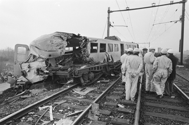 Ein Schwarz-Weiß-Bild einer Zugkollision mit einer Gruppe von Menschen in Helmen, die darum herumstehen, Strommasten, Bäume, Gebäude und einen klaren Himmel im Hintergrund.