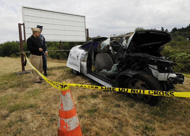 Ein Polizeibeamter in Uniform steht neben einem schwer beschädigten Auto am Straßenrand, mit eingedellter Haube und zersplitterten Fenstern, unter einem bewölkten Himmel.