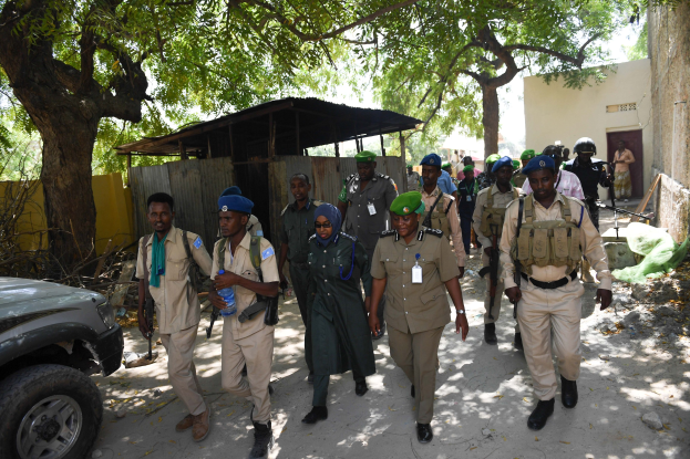 Gruppe von Polizisten in Uniform, die eine Straße entlanggehen, einige mit Waffen, mit Bäumen, Schuppen, Wänden und Gebäuden im Hintergrund.