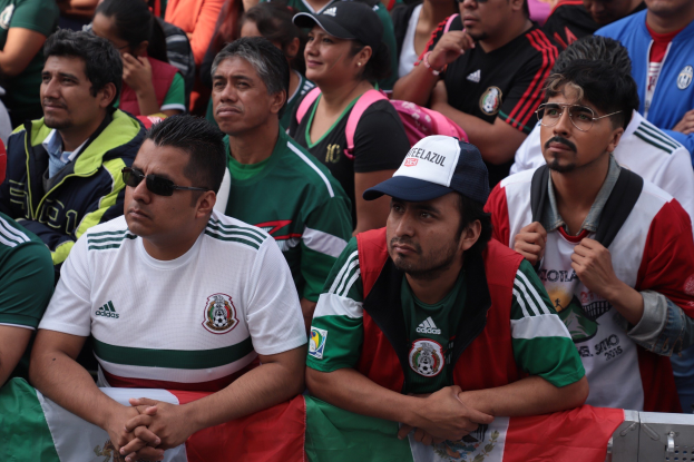 Mexikanische Fans im Stadion bei einem Fußballspiel, einige tragen Schutzbrillen, Mützen und halten Fahnen, mit einem metallischen Objekt im Vordergrund und einer Wand im Hintergrund.